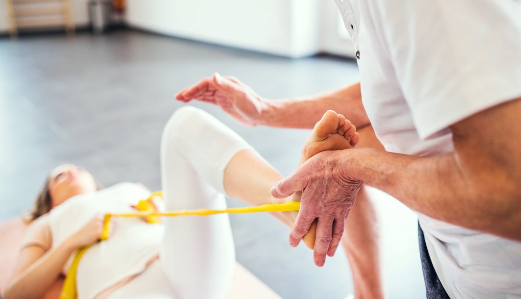 Frau liegt mit angwinkeltem Bein auf dem Boden. Ein gelbes Band umspannt das Bein. Therapeutisches Personal drückt mit der Hand gegen den Fuß.