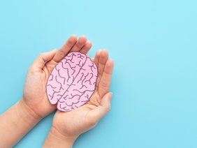 Zwei Hände halten vor hellblauem Hintergrund ein aus Papier ausgeschnittenes skizziertes Gehirn von links ins Bild.