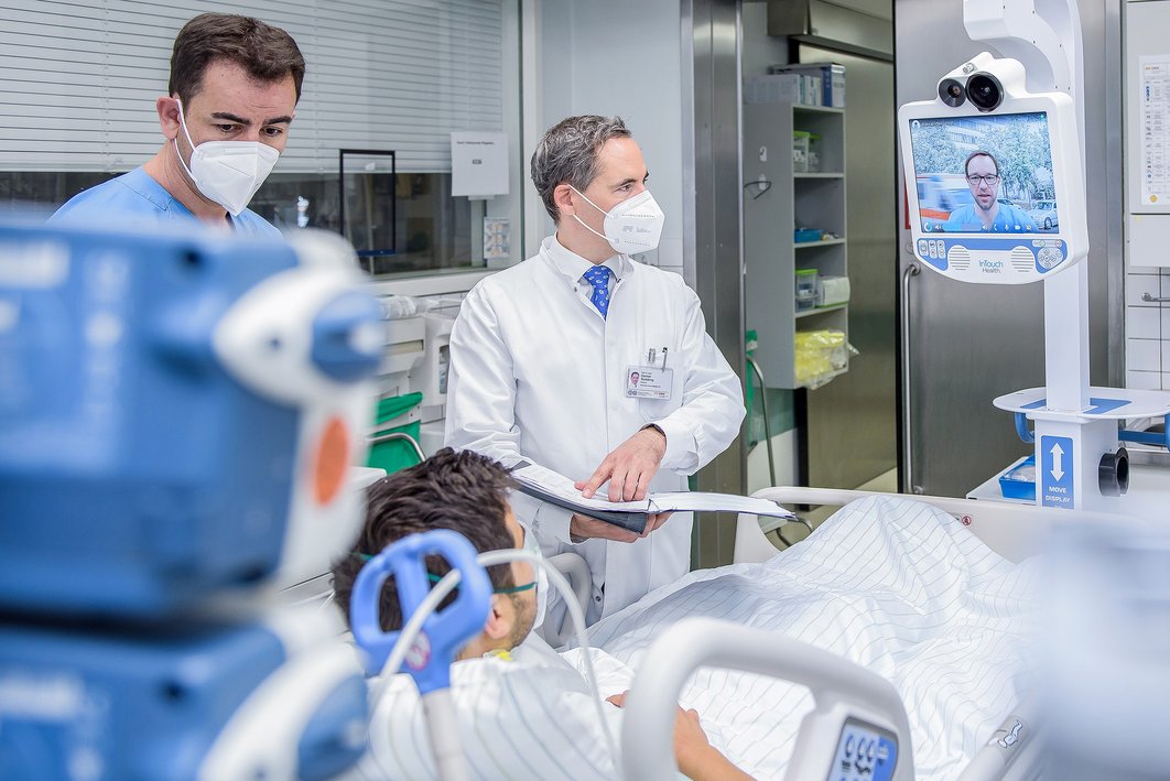 Medizinisches Gerät an einem Patientenbett im Vordergrund. Dahinter stehen zwei Männer in medizinischer Arbeitskleidung und kommunzieren über einen Visitenwagen mit Wecam mit einem anderen Arzt, der auf dem Bildschirm zu sehen ist.